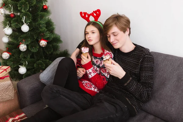 Retrato de una hermosa pareja joven sentada en el sofá en casa y usando cuernos de ciervo mientras come helado con árbol de Navidad cerca — Foto de Stock