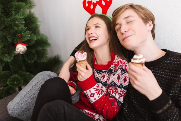 Retrato de joven pareja alegre sentada en el sofá en casa y con cuernos de ciervo mientras come helado con árbol de Navidad cerca — Foto de Stock
