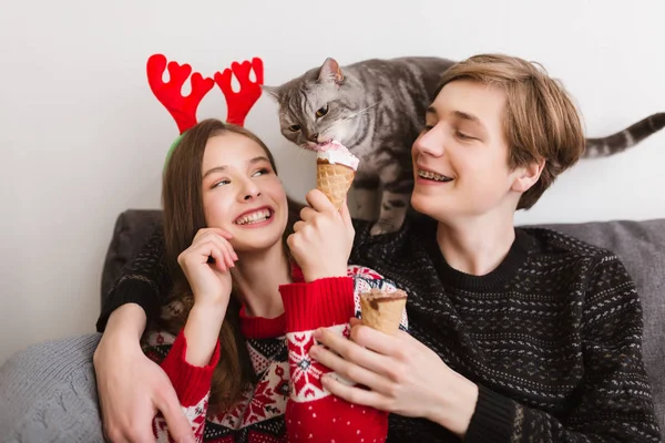 Porträt eines jungen schönen Paares, das zu Hause auf dem Sofa sitzt, Eis in der Hand und eine hübsche Katze in der Nähe, die Eis probiert — Stockfoto