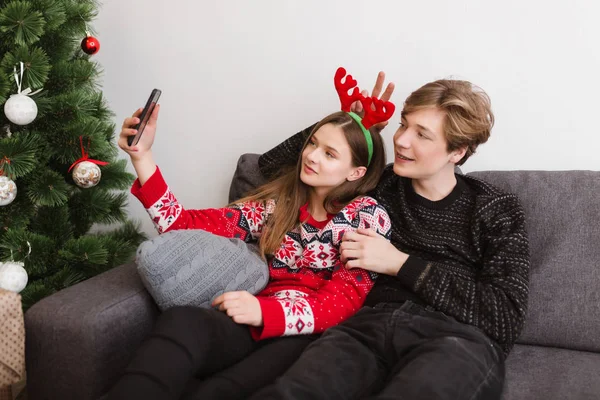 Porträt eines jungen schönen Paares, das zu Hause auf dem Sofa sitzt und zusammen mit dem Weihnachtsbaum ein Selfie macht — Stockfoto