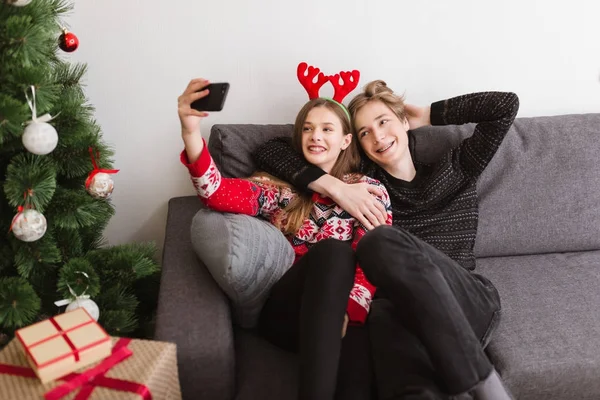 Retrato de una hermosa pareja joven sentada en el sofá en casa y felizmente tomando selfie junto con el árbol de Navidad cerca — Foto de Stock