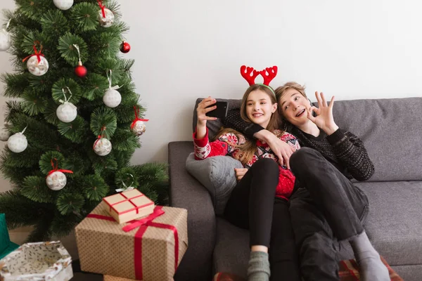 Porträt eines jungen coolen Paares, das zu Hause auf dem Sofa sitzt und fröhlich ein Selfie mit dem Weihnachtsbaum macht — Stockfoto