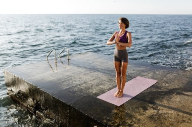 İle ayakta siyah kısa saçlı güzel kadın gözleri ve deniz kenarında meditasyon kapatın. Güzel Bayan sportif üst ve arka plan üzerinde Deniz Manzaralı yoga yaparken şort