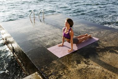 Gülümseyen kadın siyah kısa saçlı ellerinde yaslanmış ve sersem bir kenara yoga pozlar denizin kenarında eğitimi sırasında arıyorum. Sportif üst ve pratik yoga arka plan üzerinde deniz şort güzel Bayan
