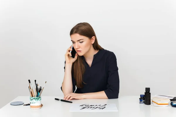 Portré, fiatal, gyönyörű lány ül a fehér íróasztal, és beszél vele mobiltelefon közben álmodozva nézett félre elszigetelt — Stock Fotó