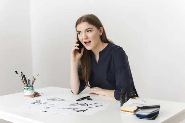 Портрет молодой красивой девушки, сидящей за белым столом и разговаривающей по мобильному телефону, счастливо смотрящей в сторону — стоковое фото