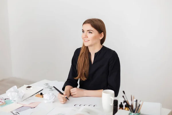 Портрет молодой улыбающейся девушки, сидящей за белым столом и пишущей заметки на бумаге, мечтательно глядя в сторону изолированной — стоковое фото