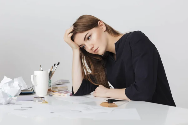Porträt einer jungen netten Dame, die am weißen Schreibtisch sitzt und den Kopf auf ihre Hand legt, während sie müde zur Seite schaut — Stockfoto