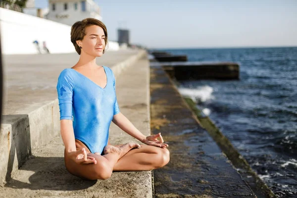 Mulher bonita sentada em pose de lótus e olhando atenciosamente em linha reta enquanto meditava junto ao mar. Retrato de senhora bonita em maiô azul praticando ioga com vista mar — Fotografia de Stock