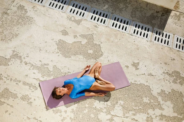 Young woman with dark sort hair lying on yoga mat and training yoga poses while thoughtfully closing her eyes. Nice lady in blue swimsuit meditating and practicing yoga isolated