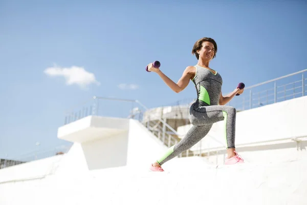 Πορτρέτο της χαρούμενη γυναίκα σε σύγχρονες αθλητικές γκρι κοστούμι και ροζ αθλητικά παπούτσια στέκεται και κάνει άσκηση με αλτήρες ευτυχώς ψάχνοντας ευθεία — Φωτογραφία Αρχείου