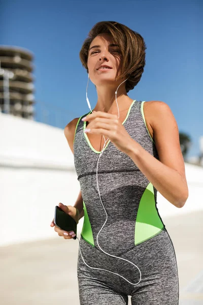 Portrait of beautiful smiling woman with brown short hair in modern gray sport suit holding cellphone with earphones while jogging isolated