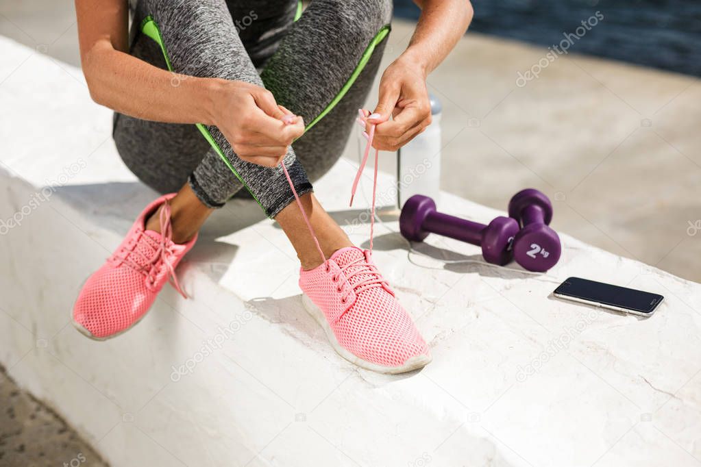 Close up photo of woman hands and legs in pink sneakers sitting and tie shoelaces with purple dumbbells, cellphone and bottle of water nearby