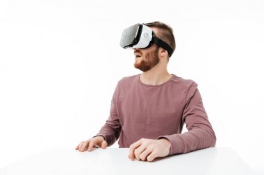 Genç gülümseyen stüdyoda oturan ve beyaz arka plan üzerinde sanal gerçeklik gözlüklü oynayan adam. Mutlu bir şekilde izole görsel gerçeklik gözlükle serin çocuk