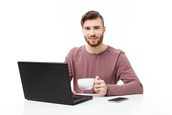 Attraktiver junger Mann, der Kaffee trinkt, am Laptop arbeitet und isoliert in die Kamera blickt — Stockfoto