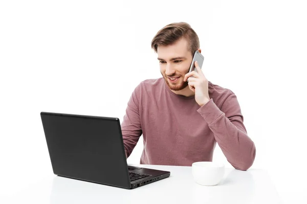 Bonito jovem trabalhando no computador portátil falando no telefone e olhando para o visor isolado — Fotografia de Stock