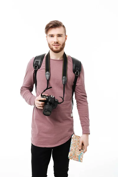 Joven hombre sonriente de pie con la cámara y el mapa en las manos sobre fondo blanco. Chico guay mirando en cámara con mochila, cámara y mapa de carreteras aislados — Foto de Stock