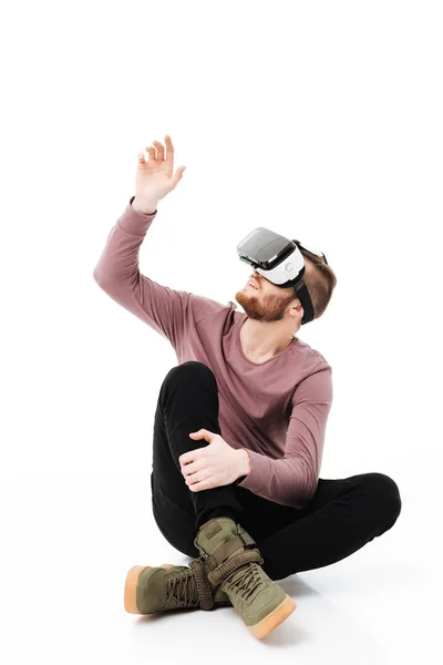 Молодий чоловік сидить у студії і грає з окулярами віртуальної реальності на білому тлі. Прохолодний усміхнений хлопчик в окулярах візуальної реальності і демонструє руку ізольовано — стокове фото