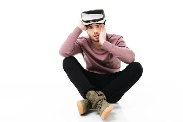 Young man sitting in studio and thoughtfully looking in camera with virtual reality glasses on head isolated. Amazed boy using visual reality glasses on white background — Stock Photo, Image