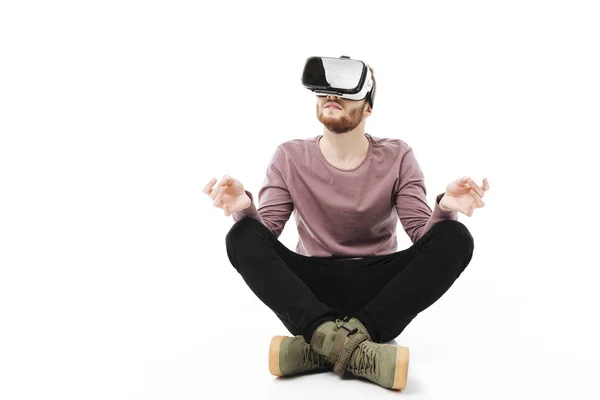 Young man sitting in studio and meditating while using virtual reality glasses isolated. Smiling boy playing with visual reality glasses on white background — Stock Photo, Image