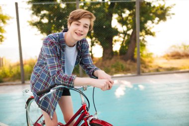Casual gömlek ayakta kırmızı bisiklet park basketbol sahası ile sarışın saçlı çocuk portresi. Mutlu bir şekilde duran klasik bisiklet ile kamera seyir gülümseyen genç adam