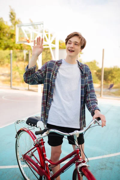 Şort ve sallayarak ve basketbol sahasına Merhaba jest duran bisiklet ile gösterilen rahat forması sarı saçlı genç adam. Çocuk holding Bisiklet gülümseyen ve mutlu bir kenara seyir portre — Stok fotoğraf