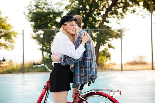 Joven hermosa chica sonriente con el pelo rubio mirando alegremente a un lado mientras abraza niño en bicicleta roja en la cancha de baloncesto en el parque. Retrato de una linda pareja abrazándose — Foto de Stock