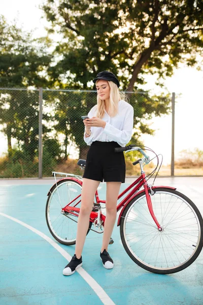 Hermosa chica con el pelo rubio en camisa y pantalones cortos de pie con bicicleta roja en la cancha de baloncesto en el parque. Jovencita en negro pico tapa de pie y el uso de teléfono celular con la bicicleta clásica cerca — Foto de Stock