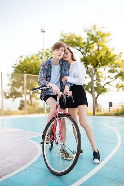 Niño en bicicleta soñando mirando en la cámara mientras hermosa chica con el pelo rubio de pie al lado y abrazándolo en el parque. Preciosa pareja joven pasar tiempo con la bicicleta en el parque —  Fotos de Stock