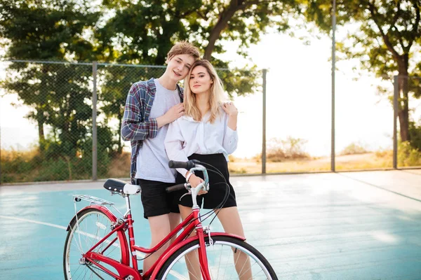 Vacker tjej med blont hår glatt tittar i kameran samtidigt som står med lycklig pojke och håller handen i park. Porträtt av söta unga par omfamnar varandra med klassiska röda cykel nära — Stockfoto