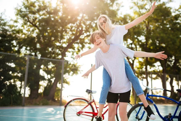 Porträtt av unga par att ha roligt tillsammans i parken med cyklar på bakgrunden. Gladlynt pojke leker med vacker flicka i solglasögon och visar plan vingar gest i park — Stockfoto