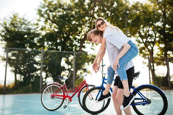 Портрет молодой пары, развлекающейся в парке с красным и синим велосипедом на заднем плане. Улыбающийся мальчик смотрит в камеру, играя с красивой девушкой в солнечных очках и держа ее на спине — стоковое фото