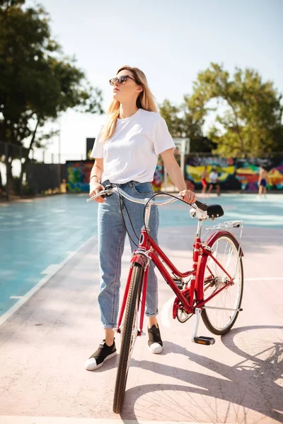 Miła dziewczyna z blond włosami w okulary, stojąc z czerwonym klasyczny rower na boisko do koszykówki w parku. Portret młodej damy przemyślane w koszulki i jeansy, Przeglądnięcie do góry trzymając rower obok — Zdjęcie stockowe
