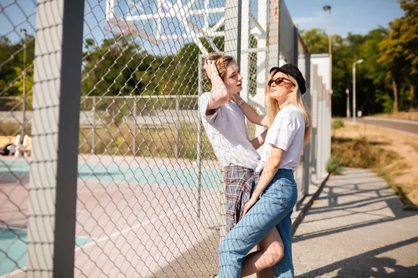 Cool jongen permanent met mooi meisje en opruimen zijn haar terwijl ze dromerig opzij kijken. Meisje in jeans en piek cap tijd doorbrengen met jongen op basketbal teller met gaas hek op achtergrond — Stockfoto