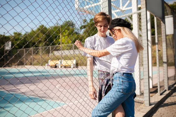 Χαμογελαστό αγόρι στέκεται με όμορφη κοπέλα και απορώντας ψάχνει στην άκρη. Όμορφο κορίτσι με τζιν και μαύρο ΚΑΠΕΛΑΚΙ με ΓΕΙΣΟ στέκεται με αγόρι στον πάγκο του μπάσκετ με ματιών φράχτη στο φόντο — Φωτογραφία Αρχείου