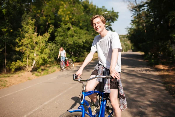 Portret młodych uśmiechnięty człowiek z blond włosami na rowerze szczęśliwie patrząc w aparacie w parku. Fajny chłopak, jazda na rowerze niebieski a spędzanie czasu w parku — Zdjęcie stockowe