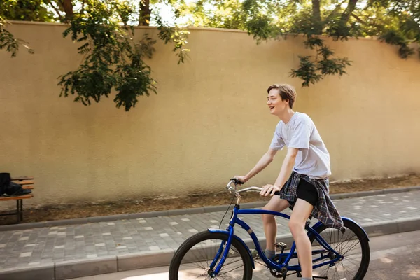 Portrait of smiling boy happily riding bicycle. Young cheerful man with blond hair having fun while riding on blue bicycle along city streets — Stock Photo, Image