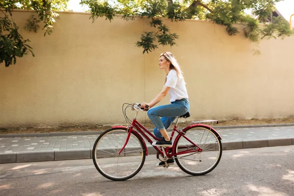 Vacker flicka med blont hår gärna rida cykel. Porträtt av ung dam i jeans och solglasögon på huvudet ha kul när du rider på röd cykel längs stadens gator — Stockfoto