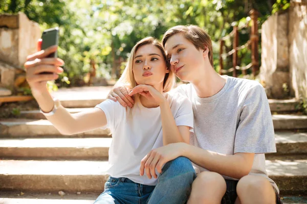 Retrato de una hermosa pareja joven sentada en las escaleras en el parque y haciendo selfie juntos. Chico fresco y chica bonita con el pelo rubio mirando en la cámara mientras toma fotos en la cámara frontal del teléfono móvil — Foto de Stock