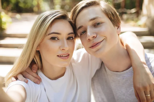 Portrait of beautiful young couple making selfie together. Cool boy and pretty girl with blond hair happily looking in camera while taking photos on mobile phone frontal camera — Stock Photo, Image