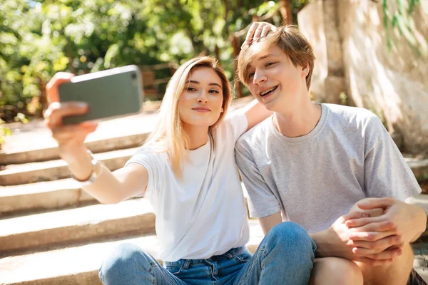 Portrét mladý pár sedí na schodech v parku a společně dělat legrační selfie. S úsměvem chlapce a krásná dívka s blond vlasy hledá ve fotoaparátu při fotografování na mobil čelní fotoaparát — Stock fotografie