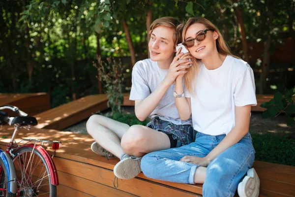 Χαρούμενο ζευγάρι κάθεται σε παγκάκι στο πάρκο και ευτυχώς ακούγοντας μουσική στο κινητό με δύο ποδήλατα γύρω από το ξενοδοχείο. Πορτρέτο των νέων χαμογελαστός άνθρωπος και όμορφη κυρία με ξανθά μαλλιά ξοδεψουν το χρόνο μαζί — Φωτογραφία Αρχείου