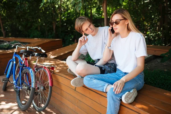 Portret van paar zitten op bankje in park met rode en blauwe fietsen in de buurt. Moe jongeman zitten en zorgvuldig kijken meisje met blond haar dat praten over haar mobiel — Stockfoto
