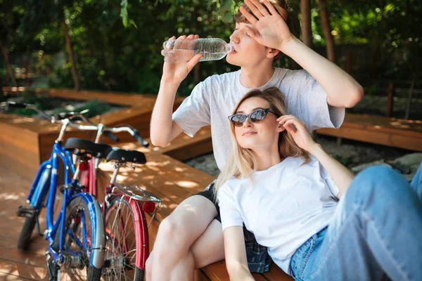 Ungt par umgås i park med två cyklar i närheten. Porträtt av pojke sitter på trä bänk och dricksvatten med vacker flicka lutande på honom och tittar glatt åt sidan — Stockfoto