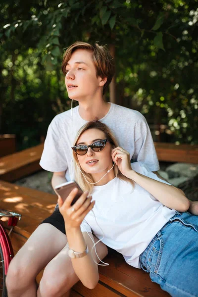Porträtt av ungt par i hörlurar lyssna musik medan du tillbringa tid i parken. Tankeväckande pojke sitter på bänken och tittar drömmande ögon medan vacker flicka lutande på honom med mobiltelefon i handen — Stockfoto