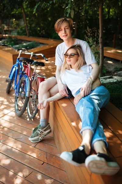 Unga söta par umgås i park med två cyklar i närheten. Porträtt av leende pojke sitter på bänken i parken och drömmande tittar i kameran medan söt tjej med blont hår lutande på honom — Stockfoto