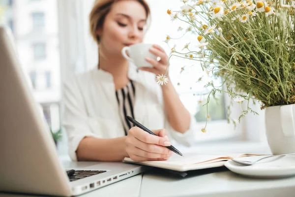 Retrato da jovem sentada no restaurante com copo na mão e escrevendo em seu caderno. Linda senhora com cabelo loiro bebendo café no café com laptop na mesa e fazendo anotações em seu caderno — Fotografia de Stock