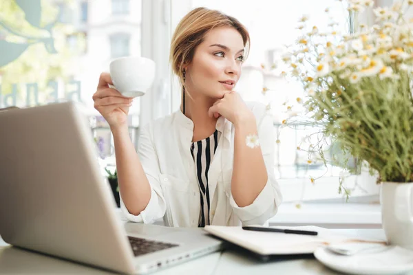 Chica joven sentada en el restaurante con taza en la mano y portátil en la mesa. Retrato de una hermosa chica con el pelo rubio descansando su barbilla en la mano cuidadosamente mirando a un lado en la cafetería —  Fotos de Stock