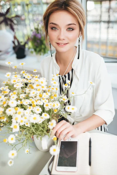 Portrait of beautiful smiling girl looking in camera with bouquet of little chamomile on table in restaurant. Pretty girl with blond hair sitting at cafe with mobile phone and notebook on tabl — Stock Photo, Image