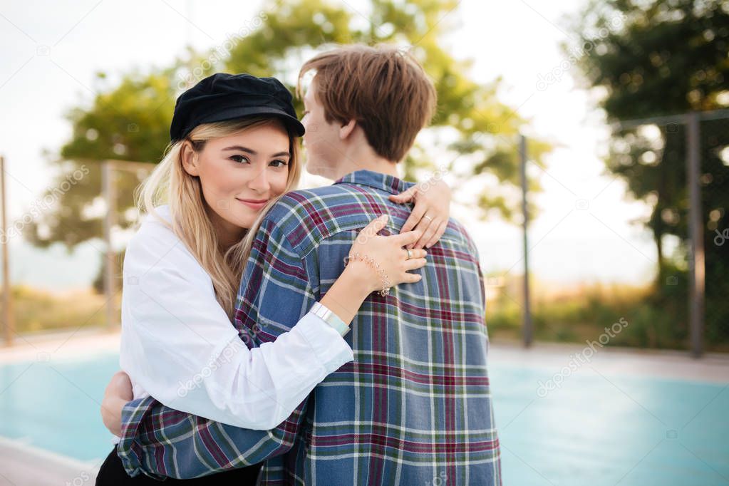 Young smiling girl with blond hair in black peaked cap happily looking in camera while embracing boy in park. Portrait of beautiful couple embracing one another